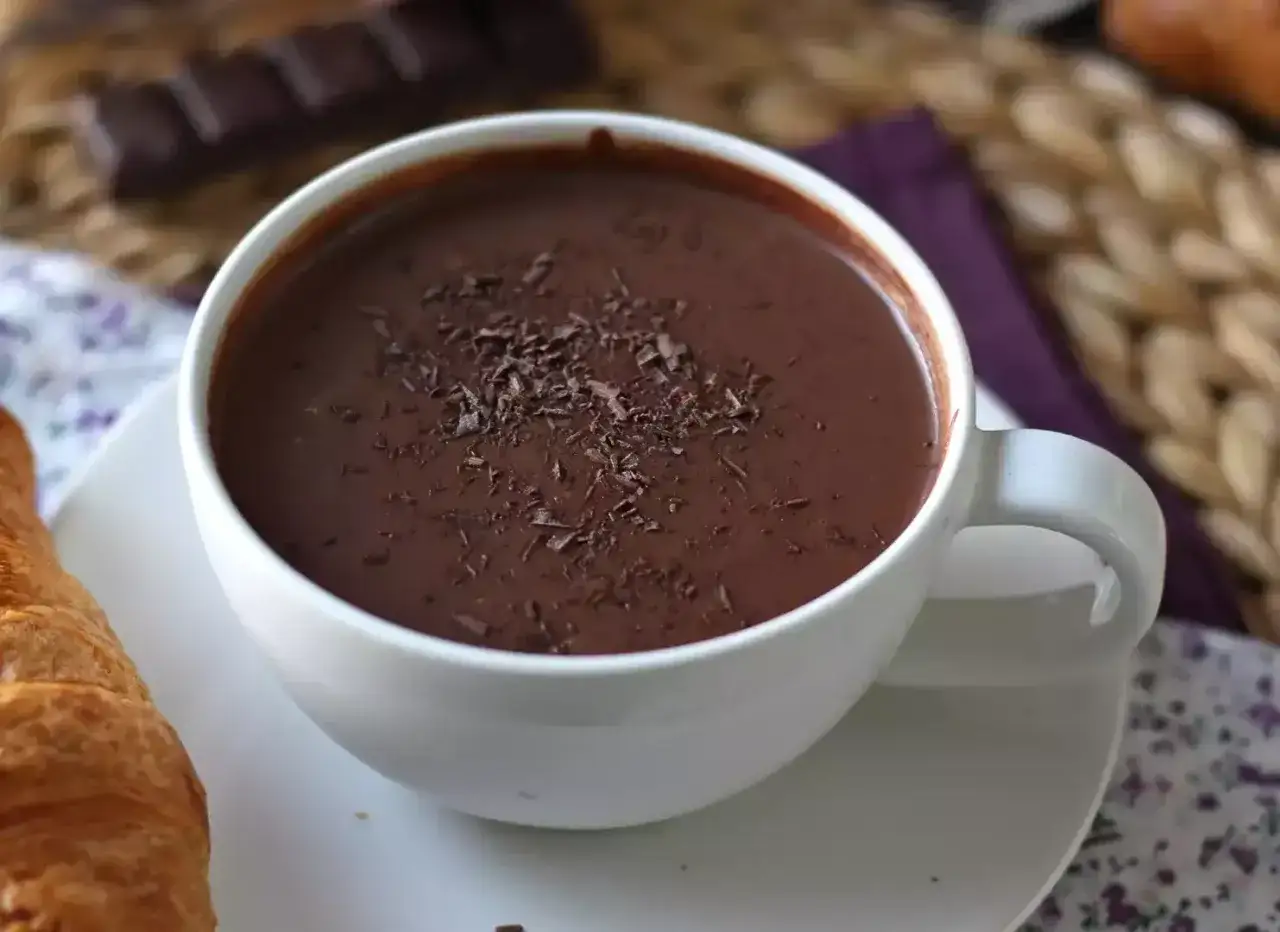 Aksamitna, gęsta czekolada w białej filiżance, posypana wiórkami czekolady. Obok croissant i tabliczka gorzkiej czekolady.